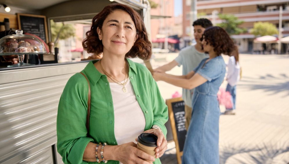 Aicha lächelt und hält einen Kaffee in der Hand, den sie gerade an einem Marktstand gekauft hat.