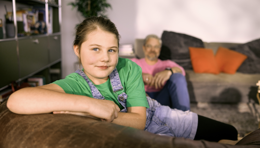 Alexa im grünen Hemd lächelt und lehnt sich an die Rückseite eines Sofas und schaut in die Kamera, während ihr Vater im Hintergrund auf dem Boden sitzt.