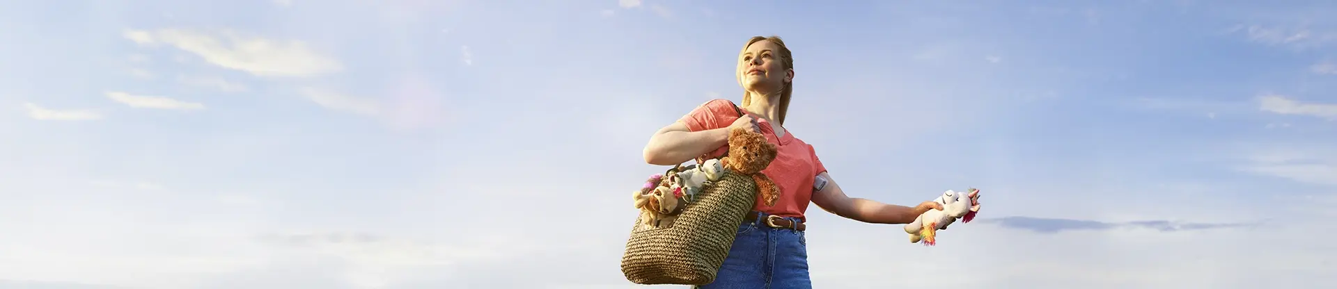 Frau steht vor blauem Himmel mit einer Tasche voller Plüschtiere und einem in der Hand.