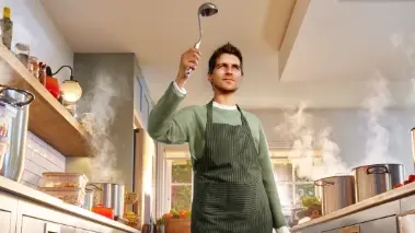 Ein Mann steht in der Küche zwischen zwei Arbeitsplatten voller Essen und hält eine Schöpfkelle hoch.