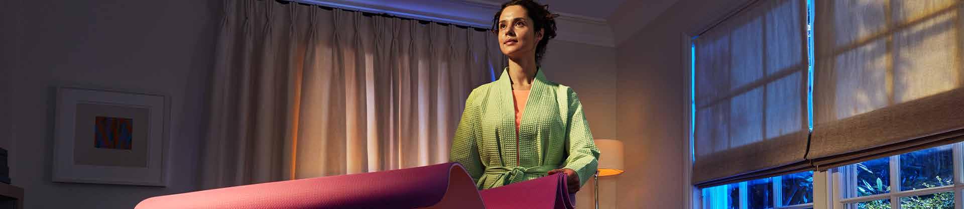 Woman holding a yoga mat indoors near a window at home.